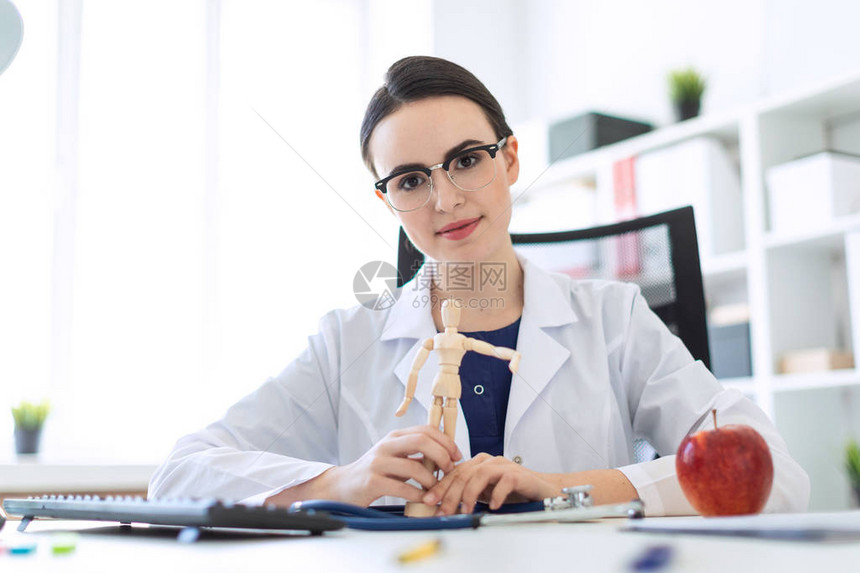 一个戴着眼镜一件蓝色衬衫和一件白色长袍的迷人年轻女孩坐在办公室里女孩面前的桌子上放着听诊器图片