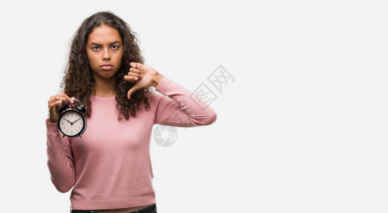 年轻的西班牙女人拿着闹钟带着愤怒的脸消极的表情示不喜欢用拇指往下伸图片