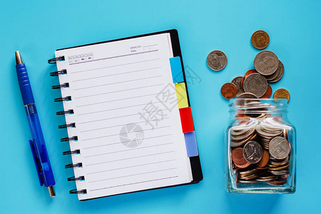 玻璃罐内外的硬币带空白螺旋笔记本和蓝色背景笔的泰币图片