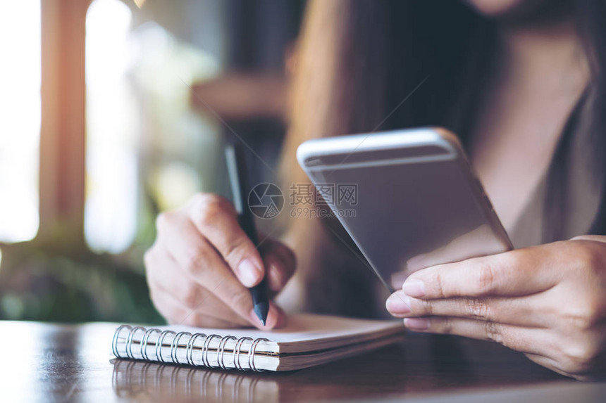 一名商业妇女在办公室使用和持有用木制表格背景的移动电话时在笔记本上写图片