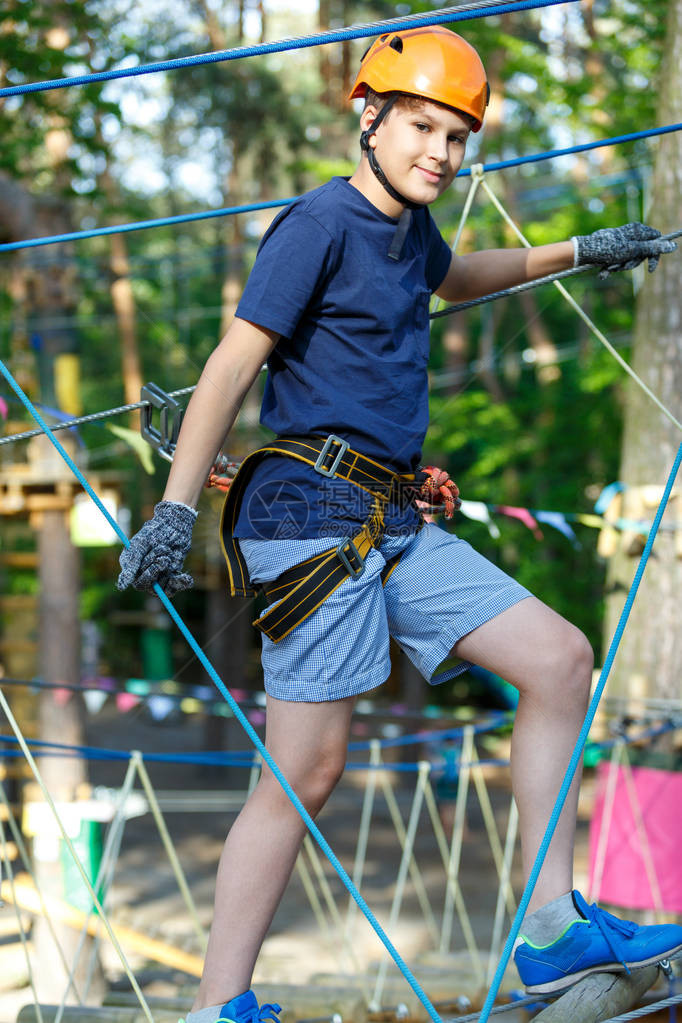 快乐可爱穿蓝T恤和橙色头盔的男孩在冒险公园玩耍玩耍握绳子爬上木楼图片