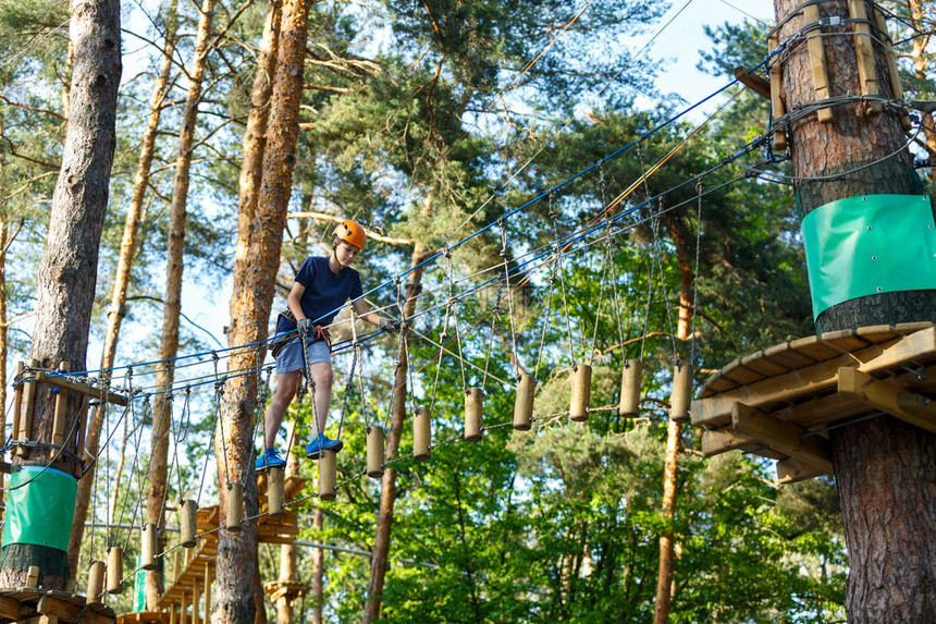 快乐可爱穿蓝T恤和橙色头盔的男孩在冒险公园玩耍玩耍握绳子爬上木楼图片