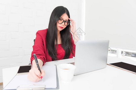 戴眼镜和穿红色西装的亚洲女商人正急忙地坐在现代办公室笔记本图片