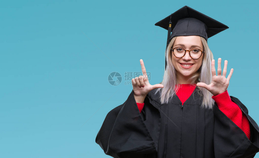 身穿毕业制服的金发年轻女郎在孤立的背景展出和指着七号手指时微笑着自信图片