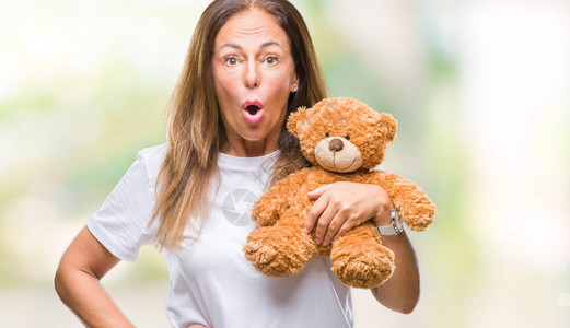 中年西班牙裔女拿着可爱的泰迪熊毛绒在孤立的背景下惊恐地惊讶地脸色惊恐图片