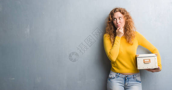 红发女青年在灰墙上拿着一箱严肃的面子认真思考问题图片