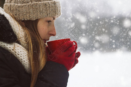 红手套女青年在雪秋天的冬季风景背上举行一个红色杯子和热茶图片