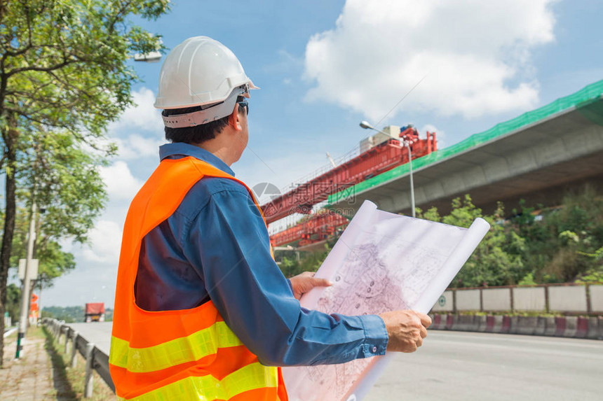 工程师或建筑师戴白色头盔在高速公路或高速公路施工现场工作或阅读施工计划作为工业或基础设图片