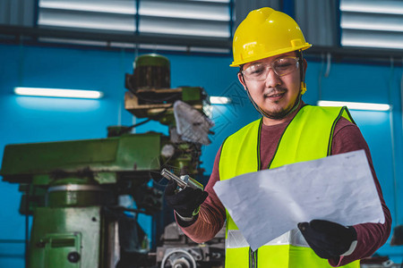 身穿安全服的亚洲机械师在金属加工厂操作专业车床前用规格纸检查设备部件图片