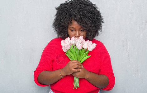 灰墙上的非洲年轻女拿着粉红花束自信地表现在聪明的面孔上认真思考图片
