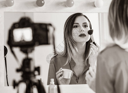 年轻caucasian博客女在视频分享网站的相机上使用化妆品在镜子图片