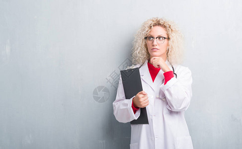 年轻金发女医生在泥黑灰墙上抱着严肃的剪贴板脸认真思考问题非图片