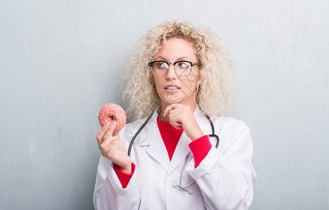 年轻的金发美食医生女人在黑灰墙上抱着甜圈的严肃面孔思考问题图片