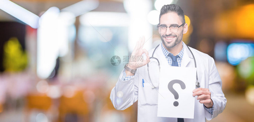 英俊的年轻医生拿着带问号的纸在孤立的背景上用手指做ok标志图片