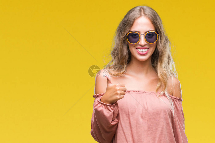美丽的年轻金发女郎戴着复古太阳镜在孤立的背景下用手做着快乐的竖起大拇指的手势批准的表情看着图片