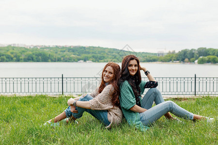 满脸笑容的黑发女孩和红色女孩坐在草地背对图片