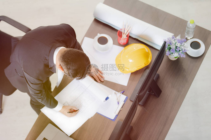 身穿黑色西装弯腰画图施工计划手拿笔黄色安全帽桌上的纸卷和文具的亚洲商人的顶视图图片