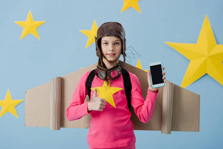 在飞行头盔中快乐的小孩拿着带空白屏幕的智能手机用蓝星背景显示拇指图片