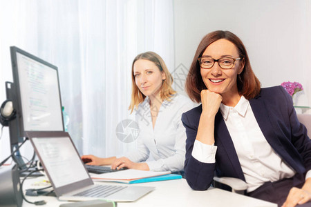坐在办公室办公桌前的快乐的中年女商人及其年轻助理的图片
