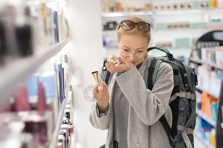 金发碧眼的年轻女旅行者穿着外套和旅行背包在机场免税店选择香水休闲女士在美容店随时地测试背景图片