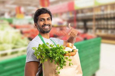 在杂货店或超市的印度男工人笑着拿蔬菜袋并用食指图片