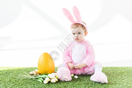 穿着有趣的兔子装可爱的婴儿坐在黄色乌龟鸡蛋旁彩色复活节鸡蛋和白边图片