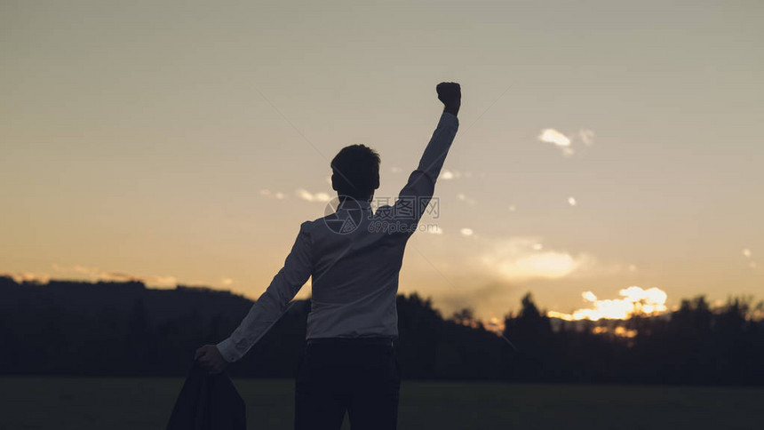 从后面看一个年轻的商人站在草原上向日出的方向转变他的手臂在胜利图片