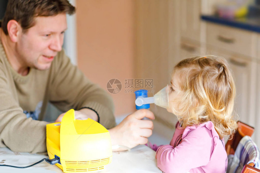 蹒跚学步的小女孩在家里用雾化器吸入父亲帮助和拿着设备孩子有流感咳嗽和支气管炎哮喘吸入器吸图片