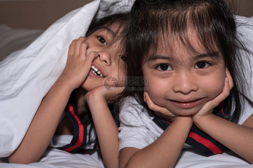 快乐的小女孩双胞胎妹在床上的毯子下玩得开心微笑和古怪床上白色毯子上的亚洲双胞