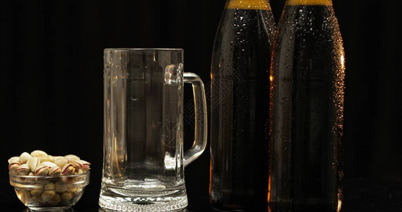 两瓶黑色背景上的冰镇啤酒一束光美丽地照亮了他们空的水杯和开心果背景图片