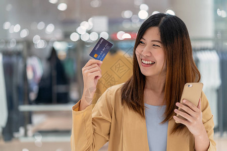 亚洲女使用带手机的信用卡在服装店背景下的百货公司网上购物技术货币钱包和在线支付概念背景图片