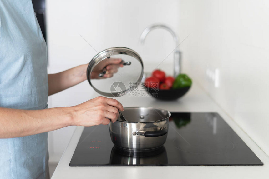 身穿蓝色围裙手拿勺子在平底锅里烹饪食物使用内置电炉站在白色现代厨房里图片