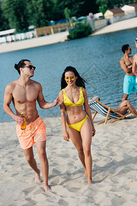 快乐的年轻男人喝着一瓶啤酒和穿着泳衣的女孩聊天在海滩图片