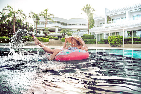 泳池边的暑假一个戴着漂亮大帽子的女人在游泳池里以甜圈的形式在一个充气圆圈上放松溅在他的脚上背景图片