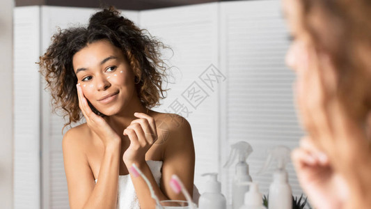 护肤理念黑色的年轻女士在浴室镜子里的眼睛下涂抹保湿霜选择图片