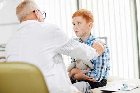 医生办公室里可爱的红发男孩在儿童诊所咨询时看高级医生的画图片