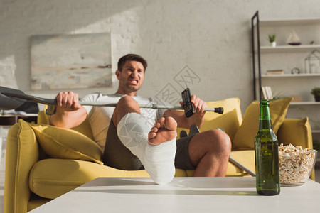 有选择的焦点愤怒的男人断腿遥控器坐在沙发上在啤酒和爆米花旁图片