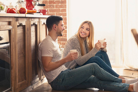 相爱的快乐小情侣愉快地交谈用茶杯聊天坐在厨房地板图片