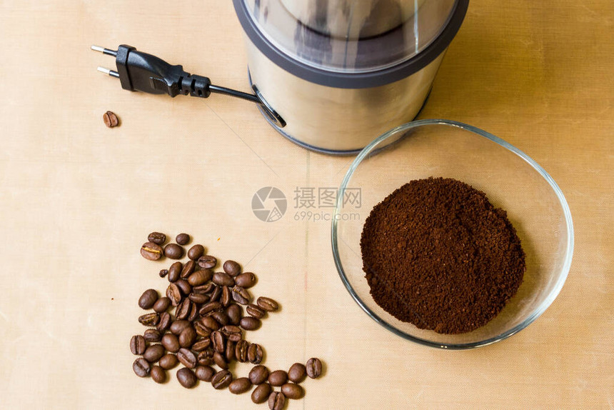 电动金属咖啡研磨机烤咖啡豆和米色桌子上的玻璃碗里的咖啡粉图片