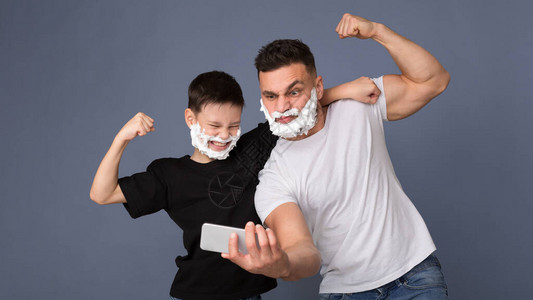强壮的爸和儿子用剃发泡沫在手机相上展示Biceps图片