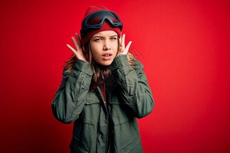 年轻的金发女孩戴着滑雪眼镜和冬季大衣在红色背景下滑雪图片