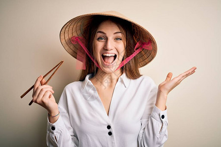 戴着亚洲传统帽子拿着木筷子的年轻漂亮红发女子非常高兴和奋图片