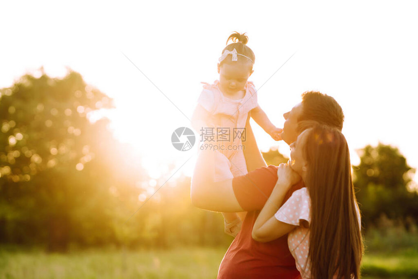 快乐的年轻家庭在日落时分在公园里散步妈爸和小女儿在夏日公园玩得开心幸福家庭的概念父母握着婴儿的手图片