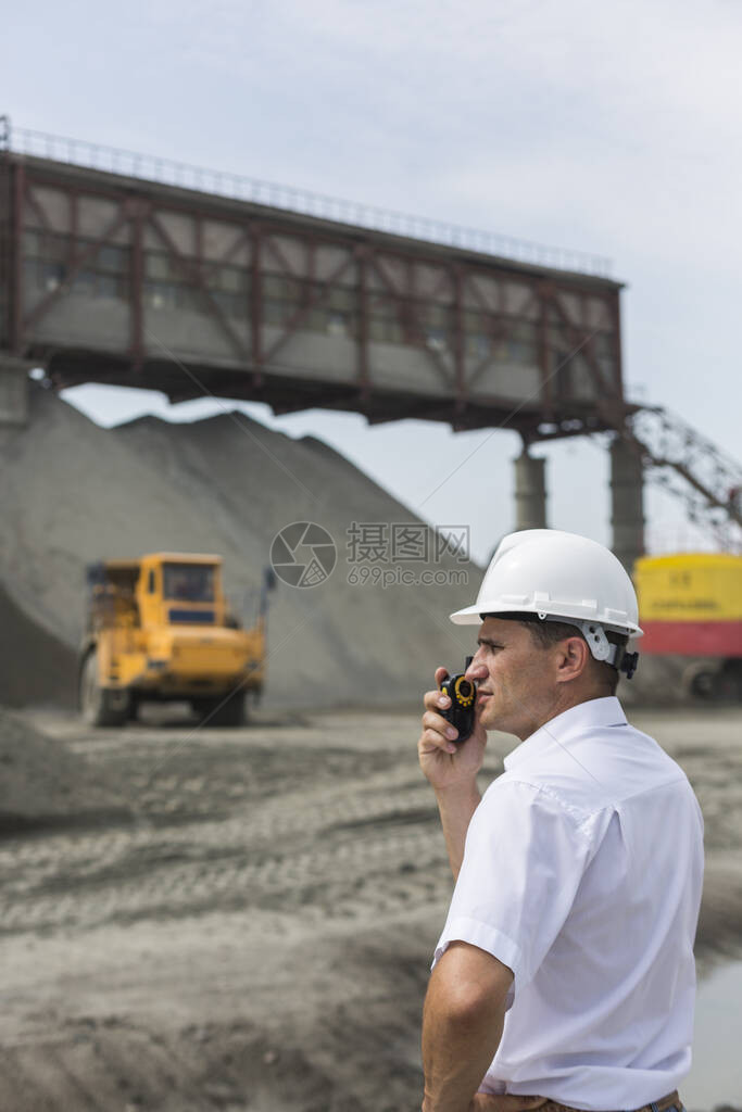 穿着白衬衫和头盔的采矿工程师拿着对讲机在采矿设备挖掘机和重型卡车中监督花岗岩加图片