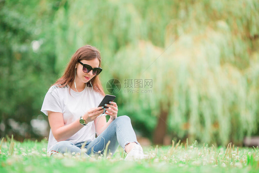 可爱的女人坐在公园里正在阅读手机上的短信美丽的女孩正在听音乐并在智能手图片