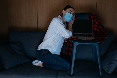 身戴医疗面具的悲伤妇女坐在笔记本电脑附近的沙发上背景图片