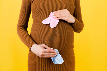 身穿棕色连衣裙的孕妇在黄色背景下将婴儿粉色和蓝色袜子靠在肚子上的特写期待双胞胎是男孩还是女孩子期待的概背景图片