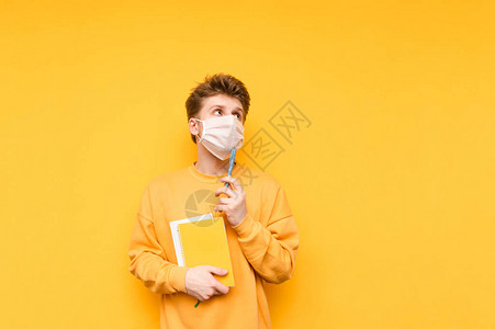 戴着医用面具手拿书和笔记本的沉思年轻男学生在黄色背景中隔开图片