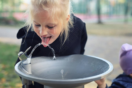 可爱的白种金发小口渴女学生在明亮炎热的夏日户外从城市公园的公共饮用水喷泉水龙头喝水儿童在背景图片