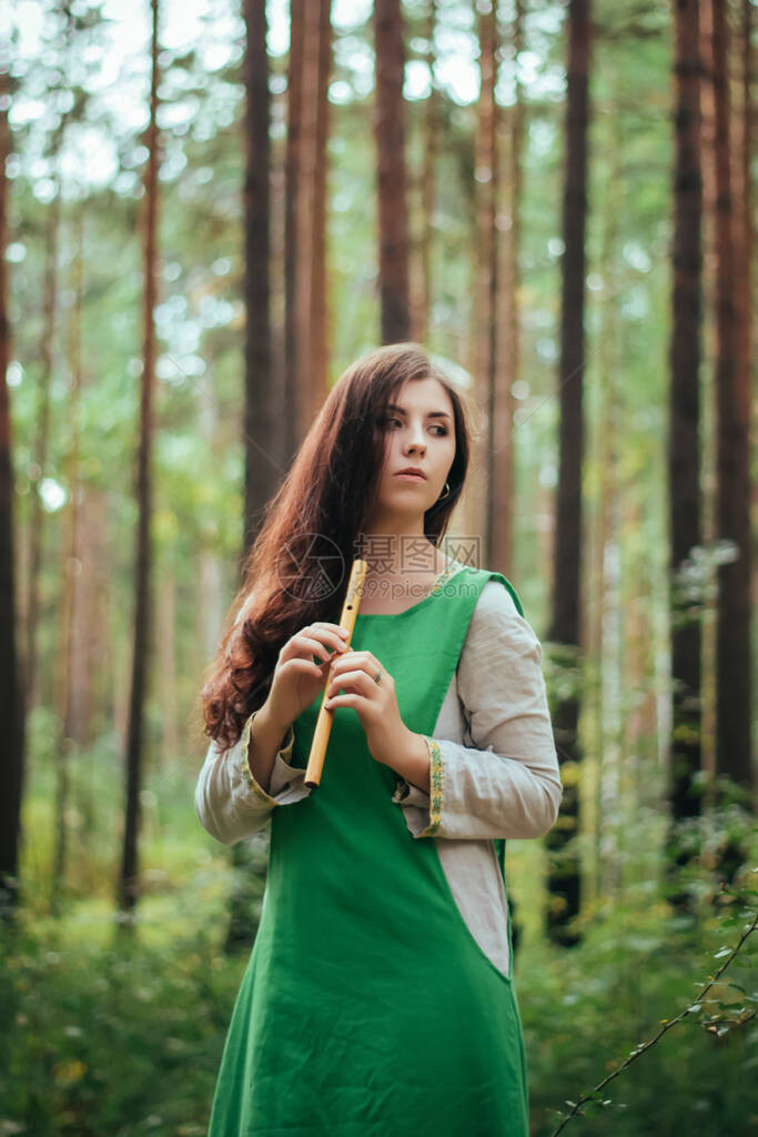 身穿绿色中世纪连衣裙的女人站在松树背景下的树林中吹奏木笛阴暗森林中的女孩在手工烟斗上演奏音乐阴暗森林图片